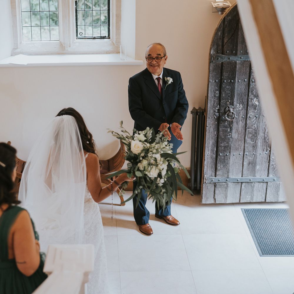 Bride in Pronovias wedding dress and veil carrying green and white rose bridal bouquet walks down stairs with bridesmaids in green bridesmaid dresses towards man in suit with white rose buttonhole before Notley Abbey wedding