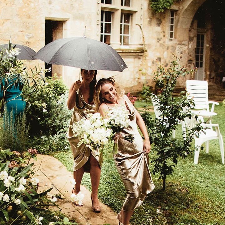 Bridesmaids walk under umbrella on wedding day whilst wearing gold silk dresses