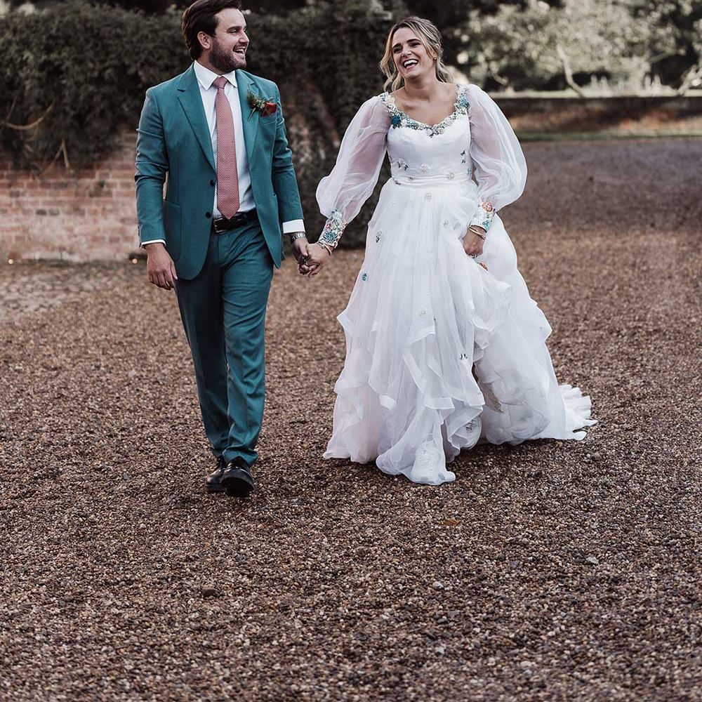Smiling groom in blue suit and pink tie holding hands with laughing bride in long mesh sleeve wedding dress walks across drive at Iscoyd Park