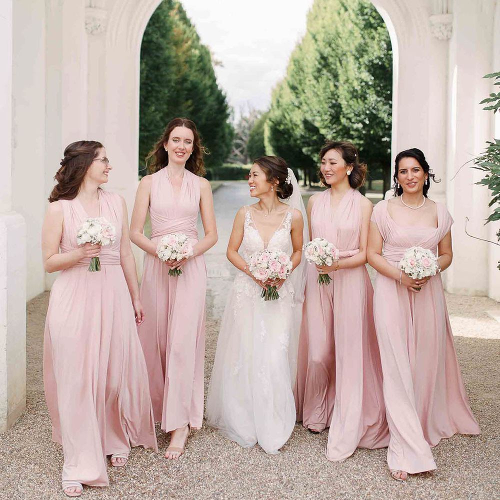 Bride walks with her bridesmaids who wear blush pink gowns with wrapped waistbands and carry white floral bouquets