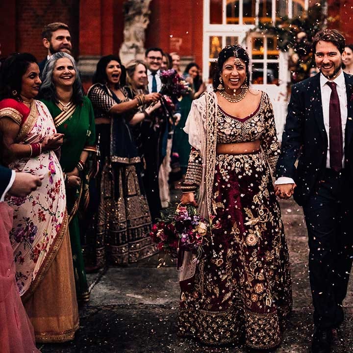 Guests cheer & throw confetti as Bride in burgundy wedding dress and Groom matches in burgundy tie 