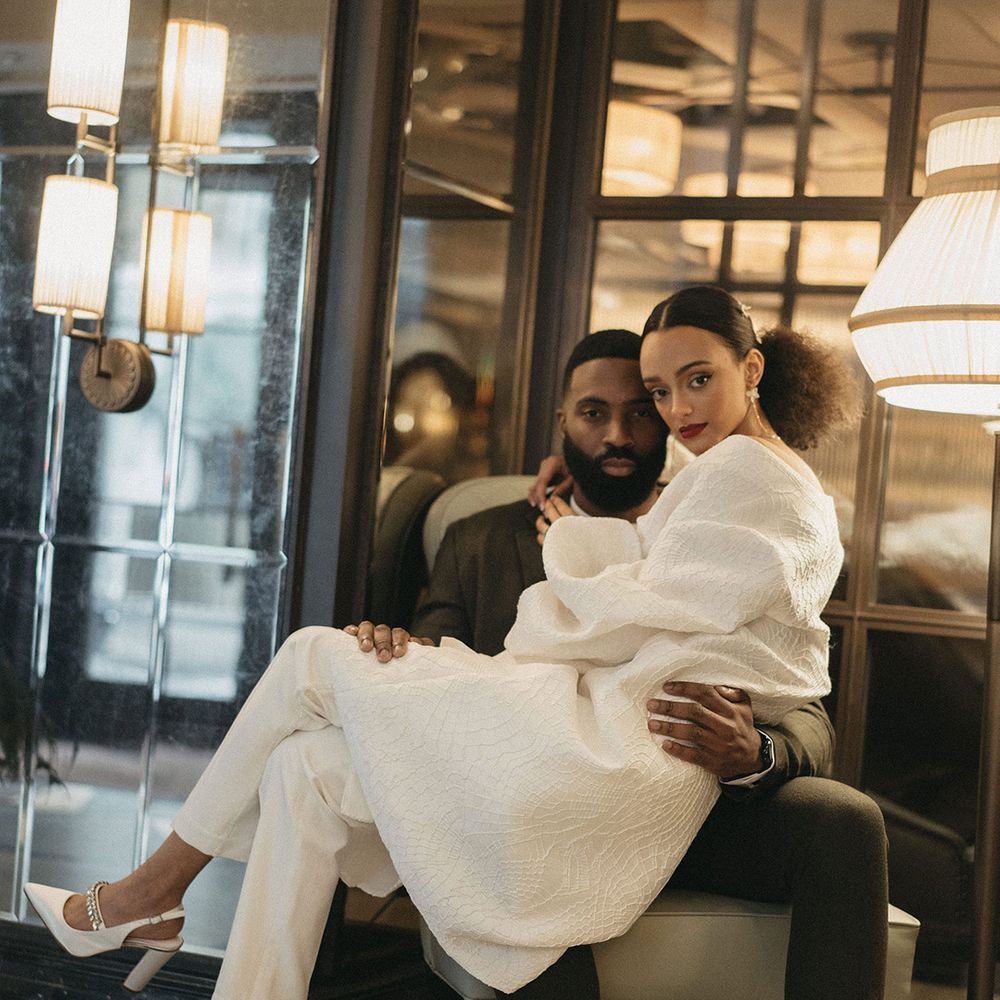 Black bride in a trouser suit and embroidered coat sitting on her grooms lap in the lobby at Mayfair Townhouse 