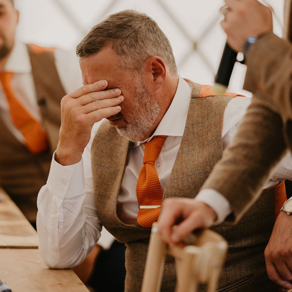 Father in matching brown waistcoat and orange tie puts his fingers to his head in embarrassment during speeches