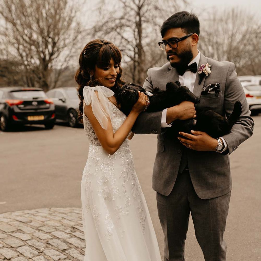 Bride in traditional white wedding dress and groom in grey suit with bow tie and boutonnière embracing black puppy