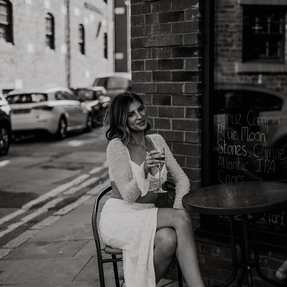 Bride in pearl embellished two piece with white feather wedding shoes holding champagne flute outside venue 