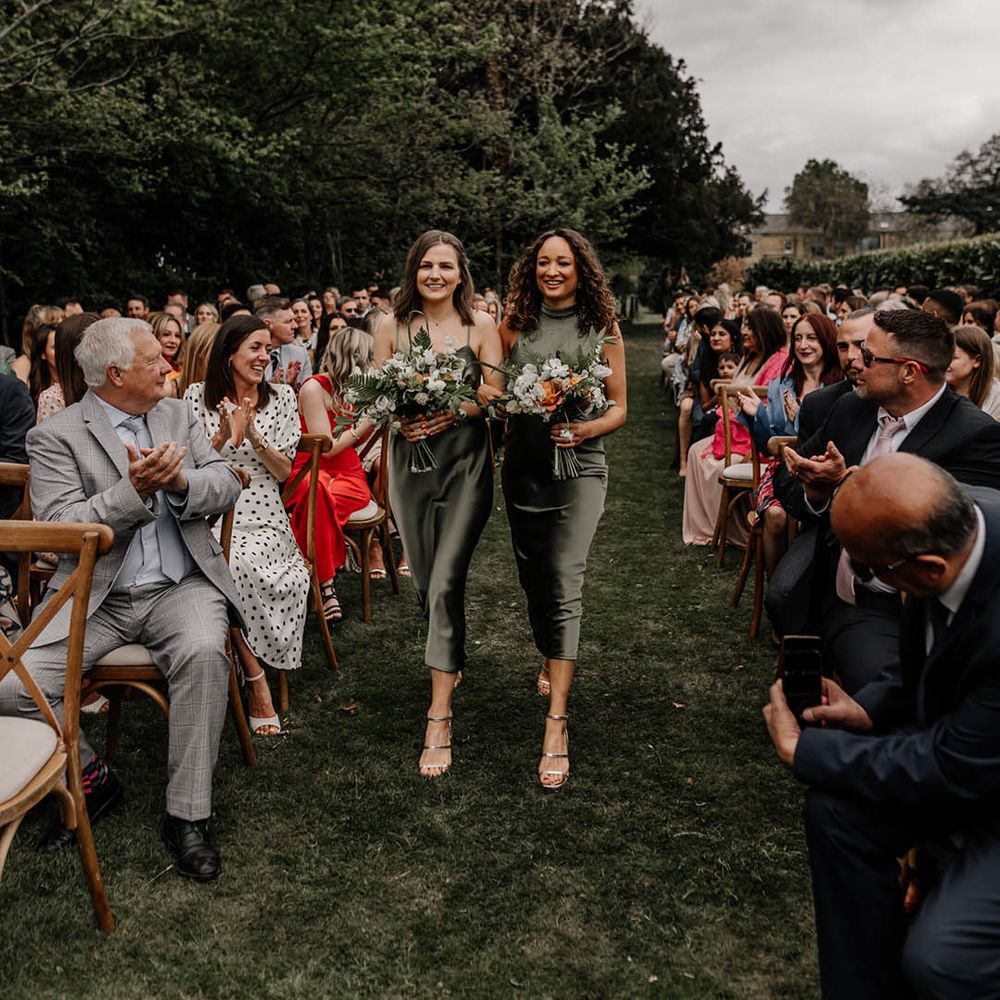Bridesmaids in satin sage green bridesmaid dresses walking down the aisle at an outdoor back garden wedding 