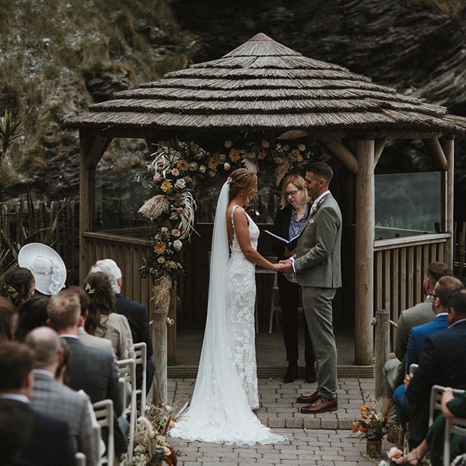 Bride & groom stand in front of beach styled hut surrounded by champagne and rust coloured florals for outdoor ceremony
