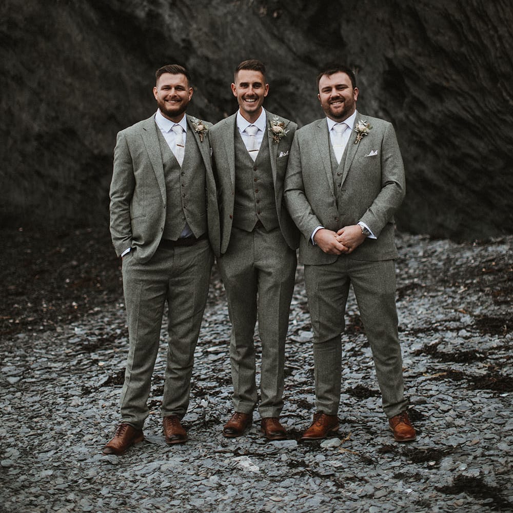 Groom stands with his groomsmen who all wear three piece suits with floral buttonholes and neutral ties 