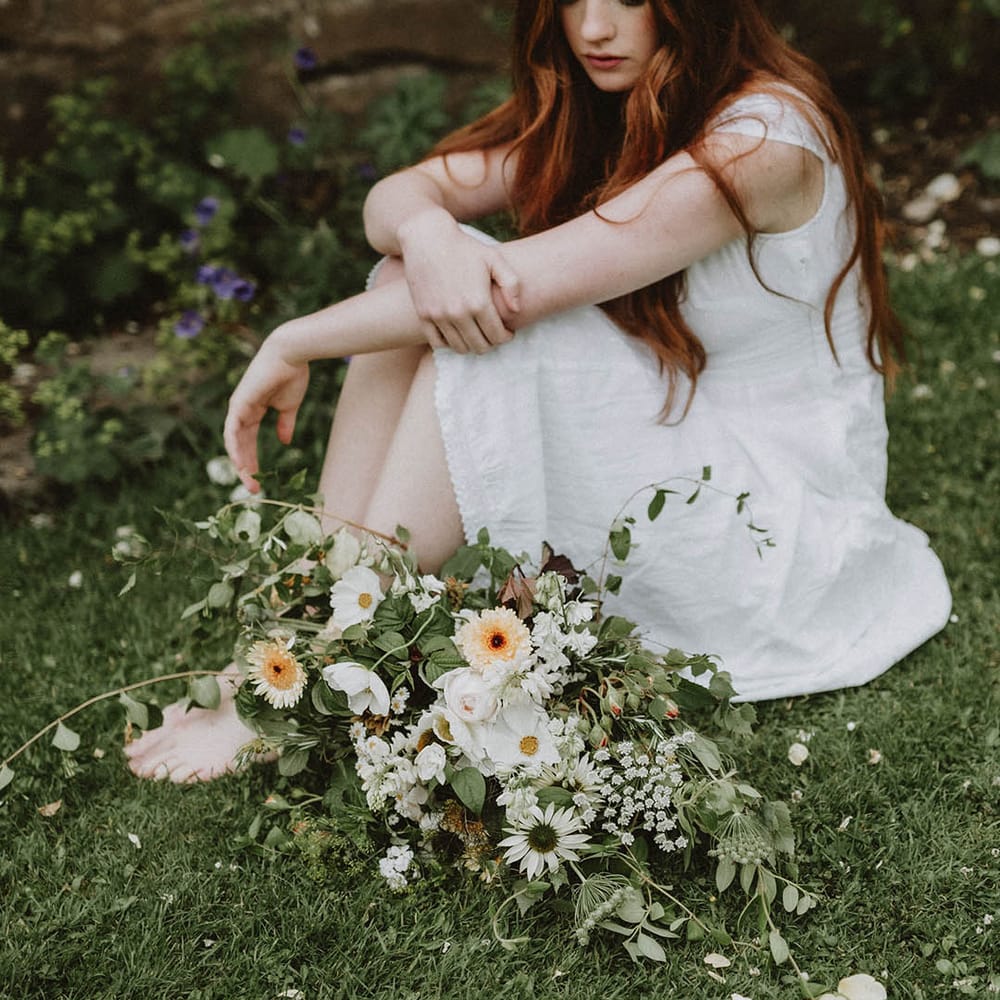 Beautiful bride with long hair and natural makeup sitting on the grass with her green, white and yellow flower and foliage wedding bouquet 