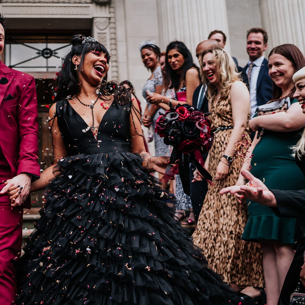 Groom wears wine red suit complete with black shirt as he walks through confetti with his bride wearing black ruffled wedding dress with V shaped neckline