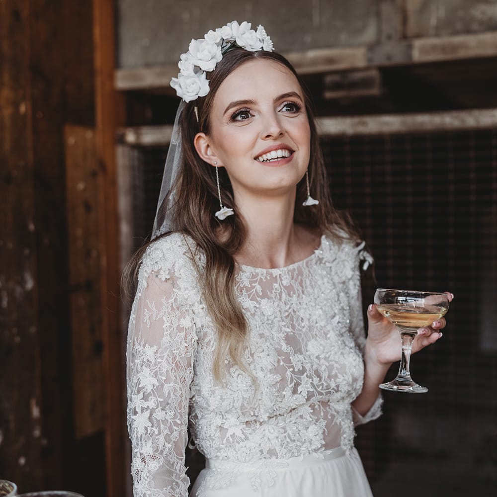 Bride in a lace wedding dress with a flower headband and flower earrings drinks champagne 