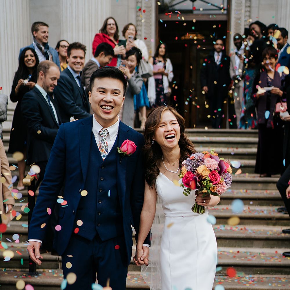Bride holds bright floral bouquet and walks with her groom through colourful confetti exit at the Old Marylebone Town Hall