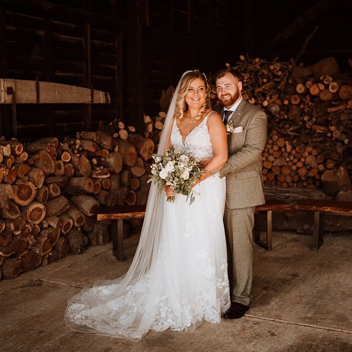 The groom in a grey tweed suit embraces the bride in a lace wedding dress from behind for their rustic wedding 