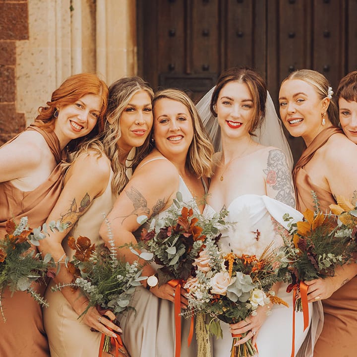 Bride with tattoos stands with her bridesmaids in Autumnal bridesmaid dresses and bouquets tied with orange silk ribbon 