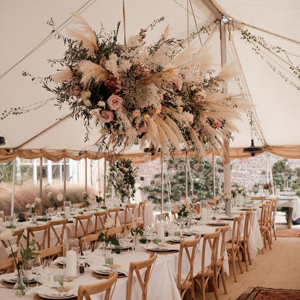 Marquee wedding reception with large flower installation with pampas grass and pink flowers over a green and white tablescape 