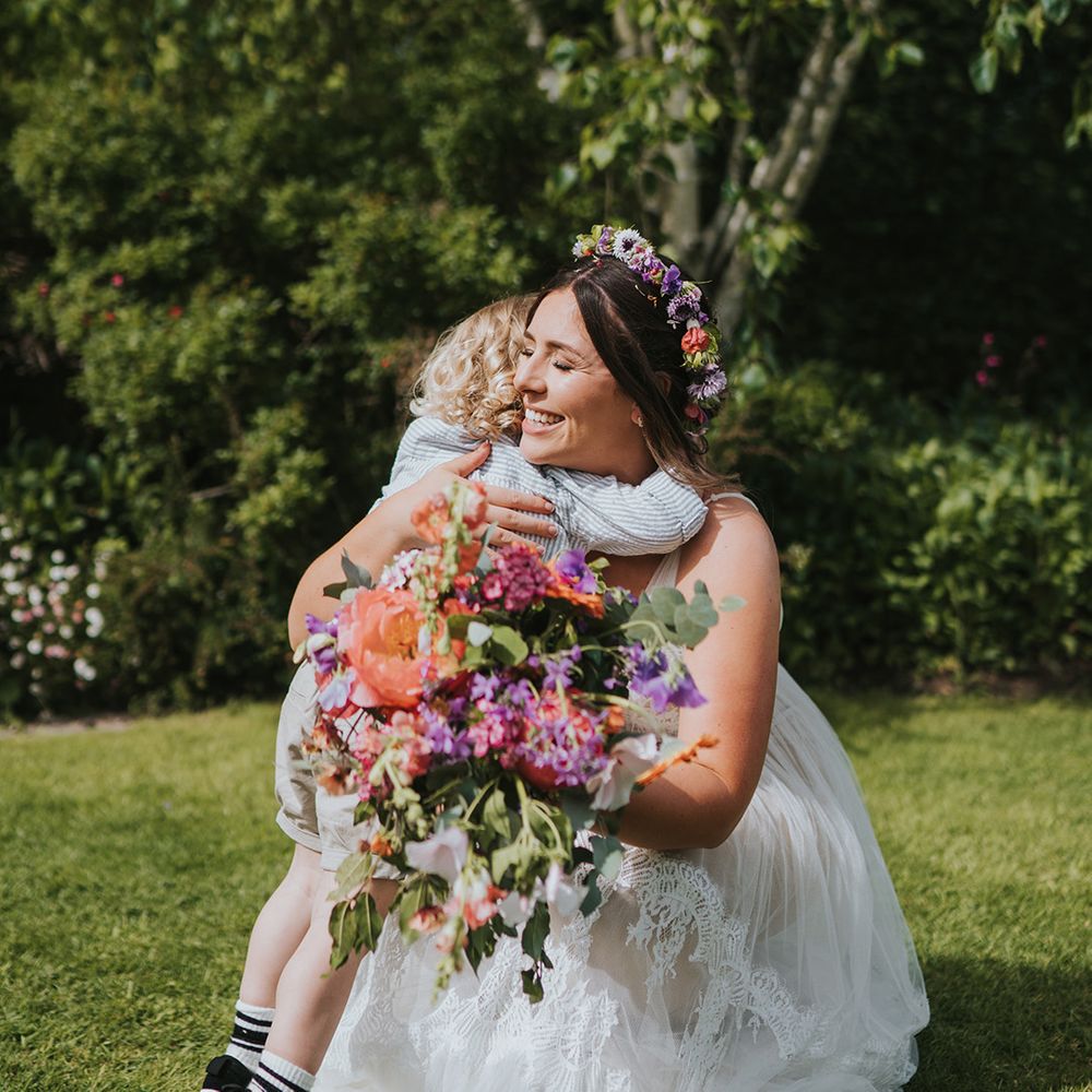 Bride holding colourful floral bouquet hugs her son outdoors
