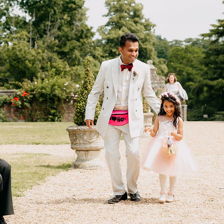 Flower girl in pale pink tulle dress walking with a groomsman in a white suit and neon pink fanny pack full of confetti 