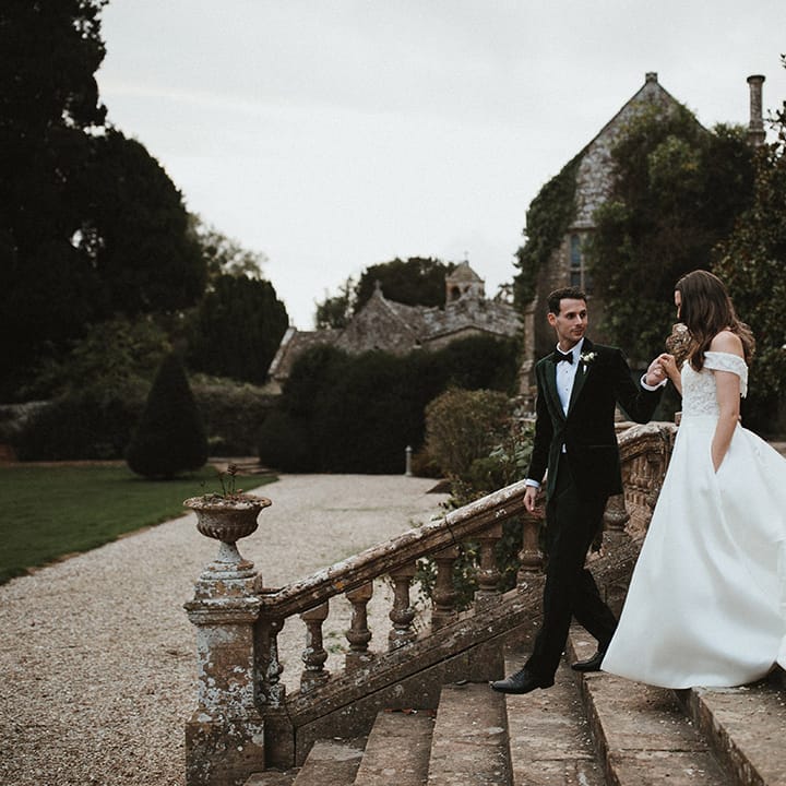 Groom in dark green velvet tuxedo walking down the stairs with the bride in an off the shoulder lace wedding dress 