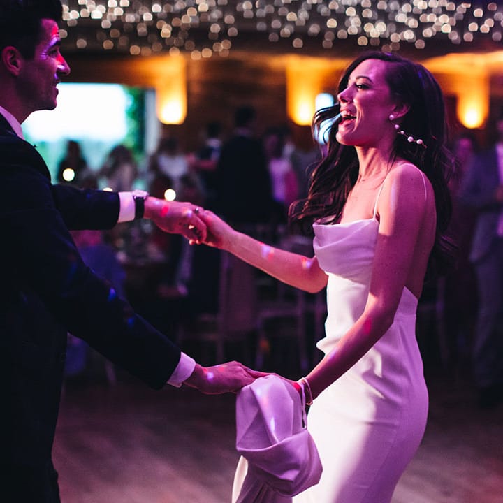 Groom in dark navy suit sharing the first dance with the bride in a satin gown as pink lights shine on them 