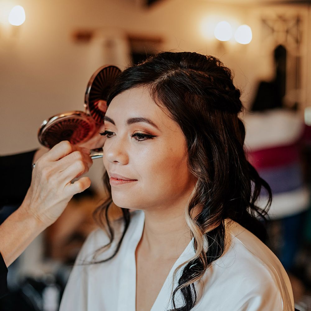 Bride sits in a white robe getting her makeup done for the wedding day with her hair curled in a half up half down hairstyle 