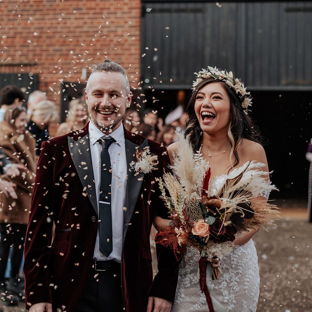 Groom in velvet suit jacket laughing as he walks along with the bride in an off the shoulder lace wedding dress with boho flower crown having confetti exit 