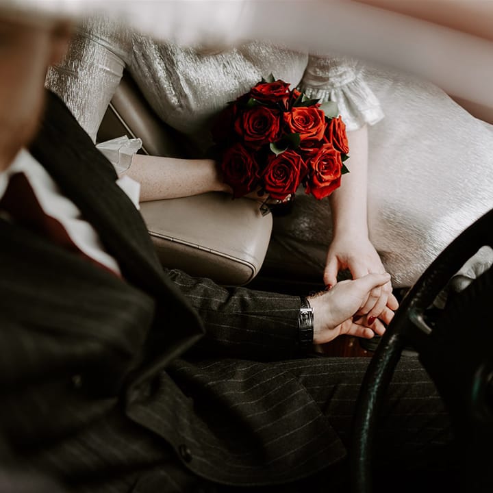 The groom in a pinstriped wedding suit driving the wedding car holding hands with the bride in a metallic wedding dress holding the red rose bouquet 