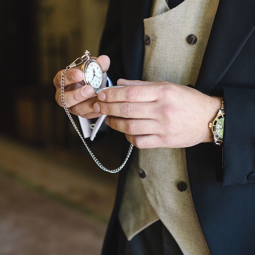 Groom in navy suit with beige waistcoat holding silver pocket watch groom accessory for the wedding