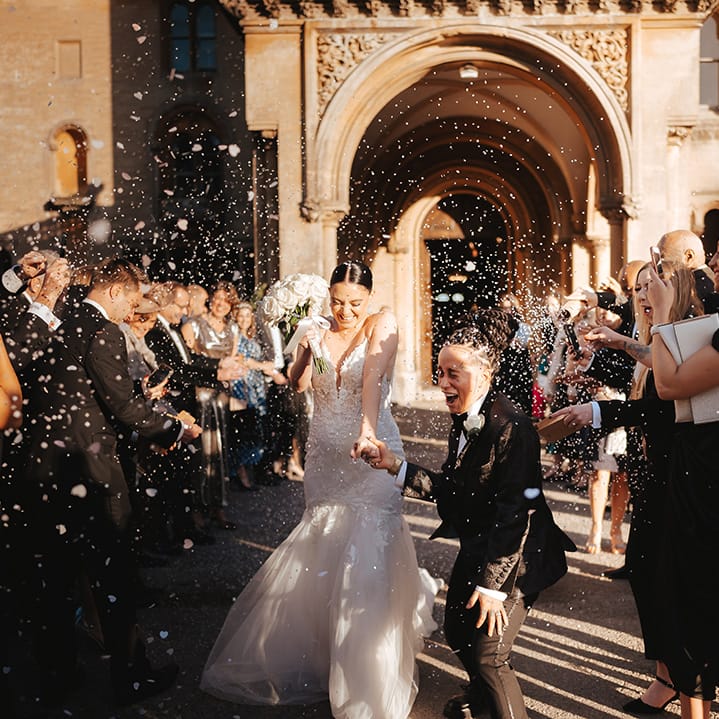 Bride in lace sleeveless v neck Enzoani wedding dress with tulle finish and church length veil holding hands with Bride in black snakeskin print tuxedo with black bowtie and white rose boutonniere holding white garden rose bridal bouquet doing white petal confetti exit at wedding