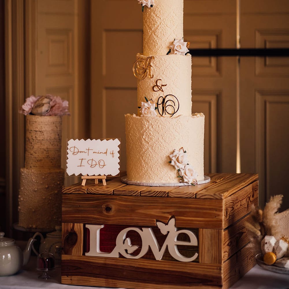 Three tier buttercream frosted wedding cake with delicate pattern, iced pink roses and personalised gold lettering 