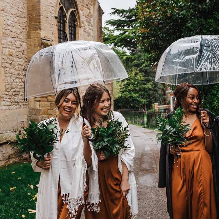 Bridesmaids in strappy sleeveless front slit burnt orange bridesmaid dresses holding foliage bouquets and shawls standing underneath umbrellas outside of church wedding