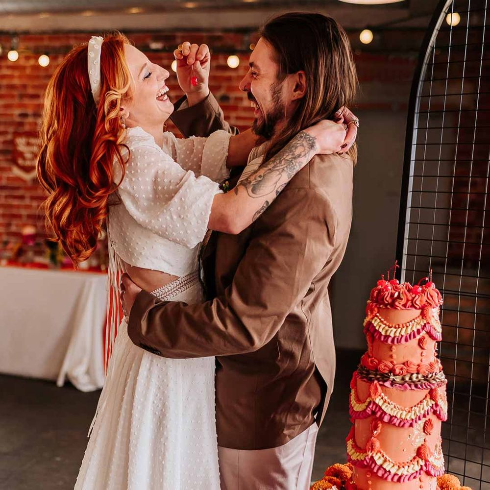 Groom in brown grooms blazer feeding bride maraschino cherry in cut out back detail short sleeve wedding dress standing by retro wedding cake at Northern Monk Refectory Leeds