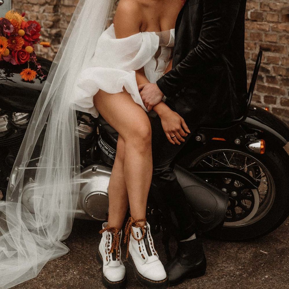 Groom in wet look black grooms suit with leopard print shirt and pink and orange boutonniere standing with bride in off shoulder tulle short wedding dress with church length veil and white platform Dr Martens wedding shoes