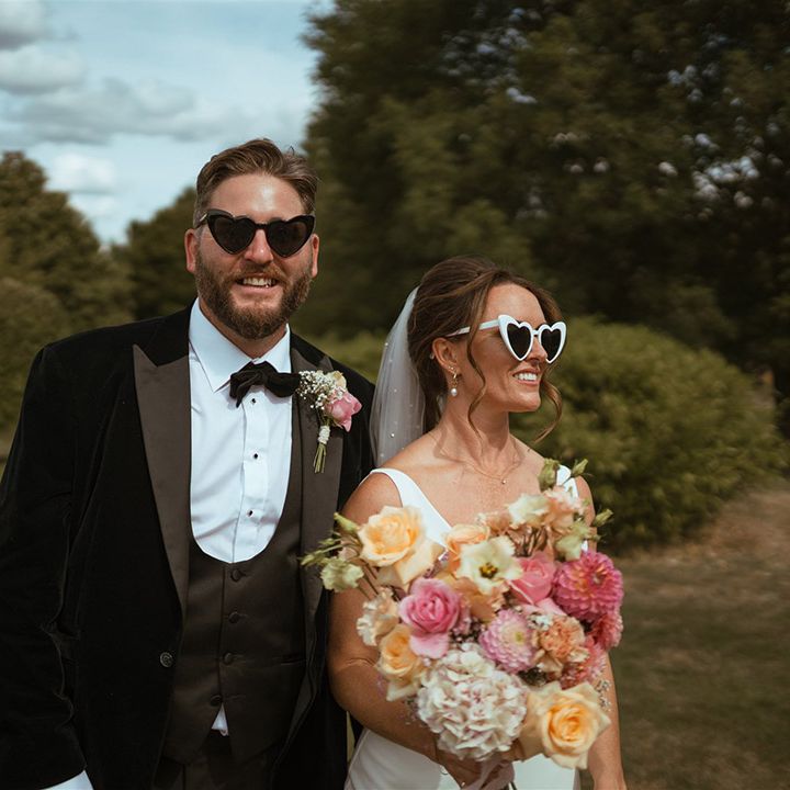 Bride in white wedding satin slip dress with colourful bouquet and retro white sunglasses with groom in black tuxedo with sunglasses