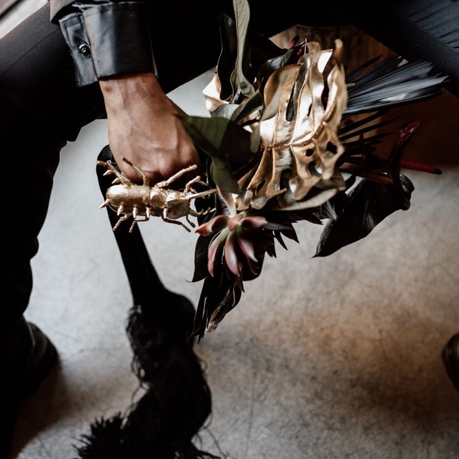 Groom holds bouquet with gold accent colour and gold beetle accessory 