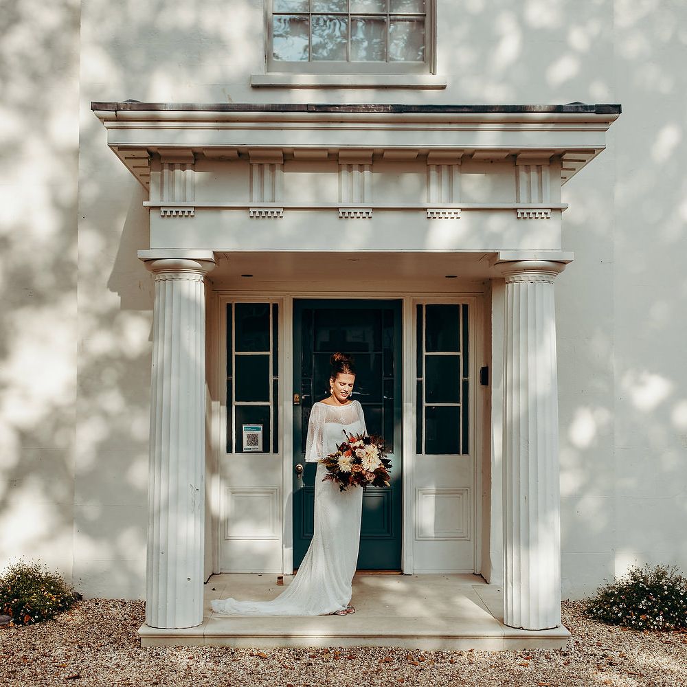 Bride with brown hair and red lipstick stands with bouquet in front of white house