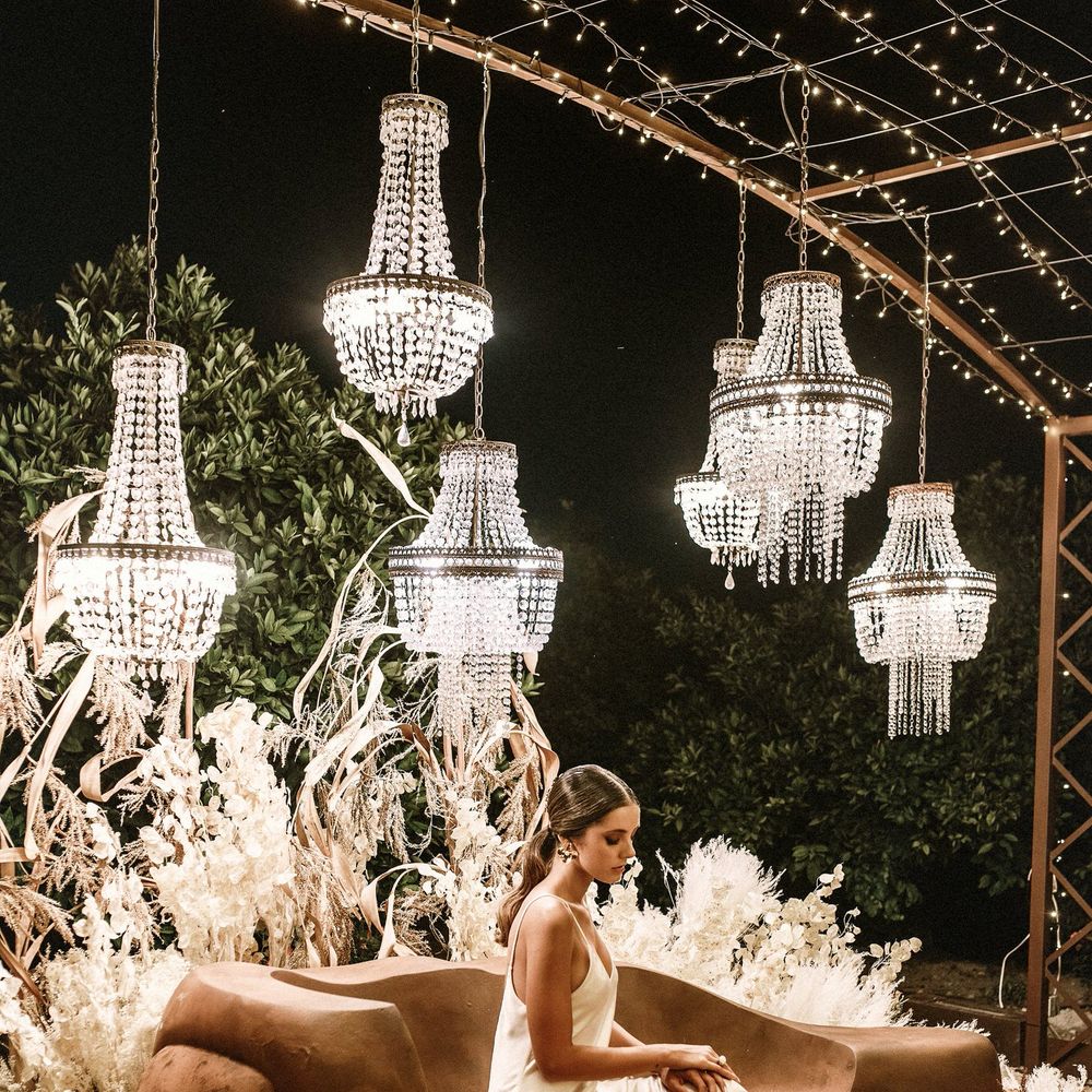 Bride in a satin slip wedding dress sitting under a canopy of lights and chandeliers with a floor full of wicker rugs 