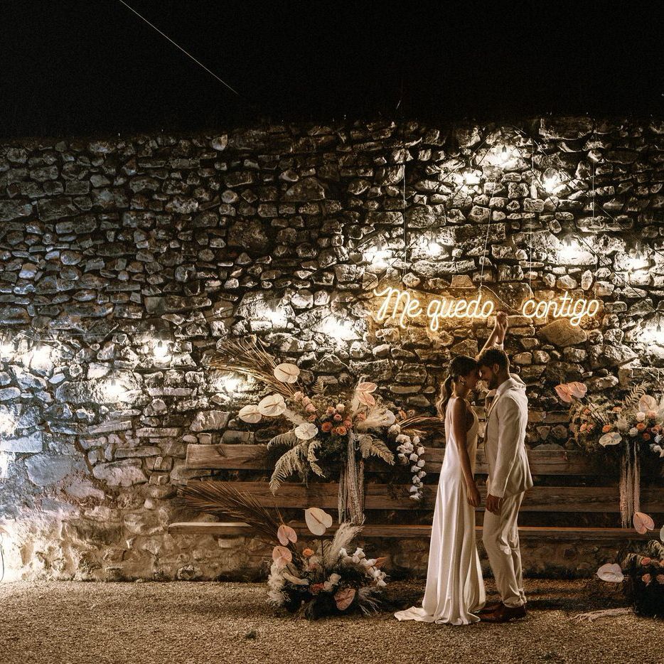 Outdoor seating area with the bride and groom standing in front of an exposed brick wall decorated with a neon sign, festoon lights and boho wedding flowers