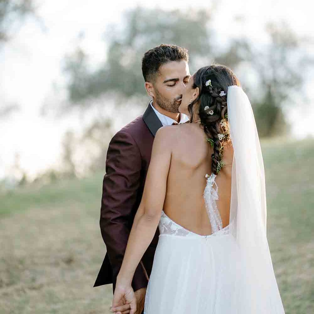 Bride clutches large bouquet wearing backless wedding dress with groom in maroon suit