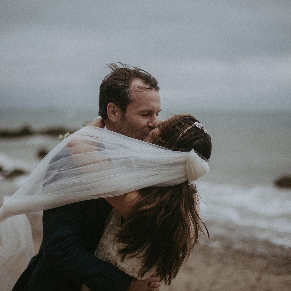 Bride & groom embrace with the sea in the background and brides veil blows around them