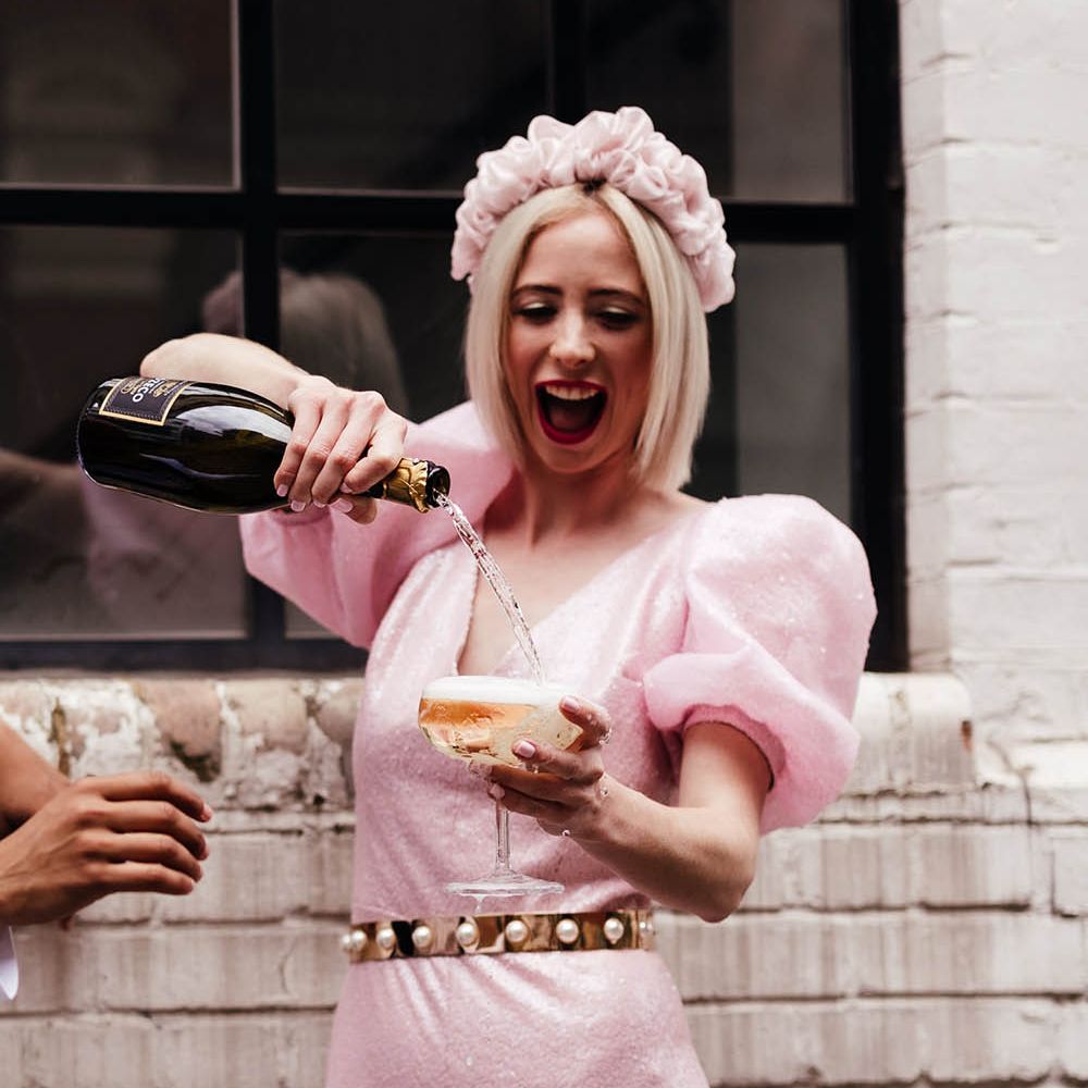 Bride in a custom-made pink jumpsuit and appliqué headband pouring champagne