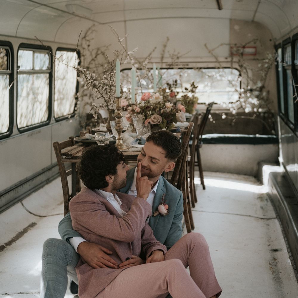 Grooms sit beside wooden table in rustic setting with pastel floral decoration and brass candle holders  