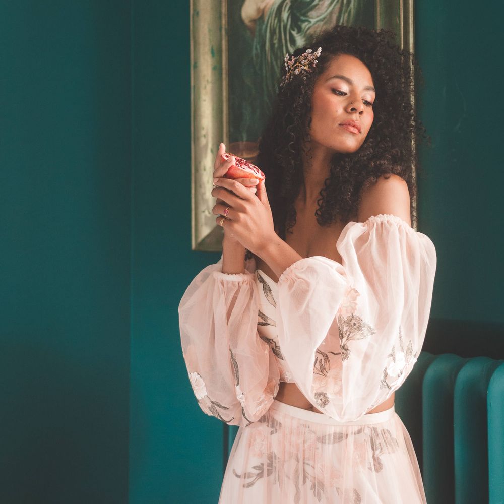 Black bride with long curly hair in blush pink bridal separates holding a pomegranate