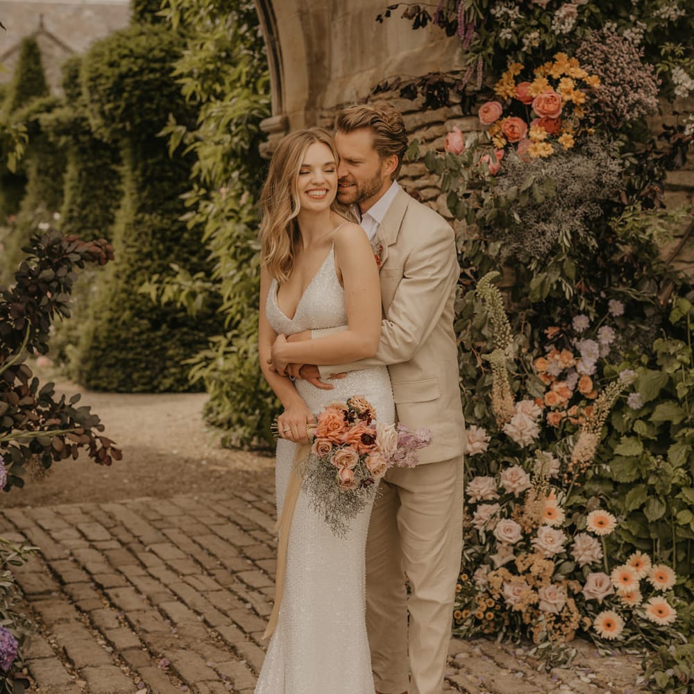 Groom in a beige suit embracing his bride in a sequin Made With Love wedding dress by the castle arch at Euridge Manor 