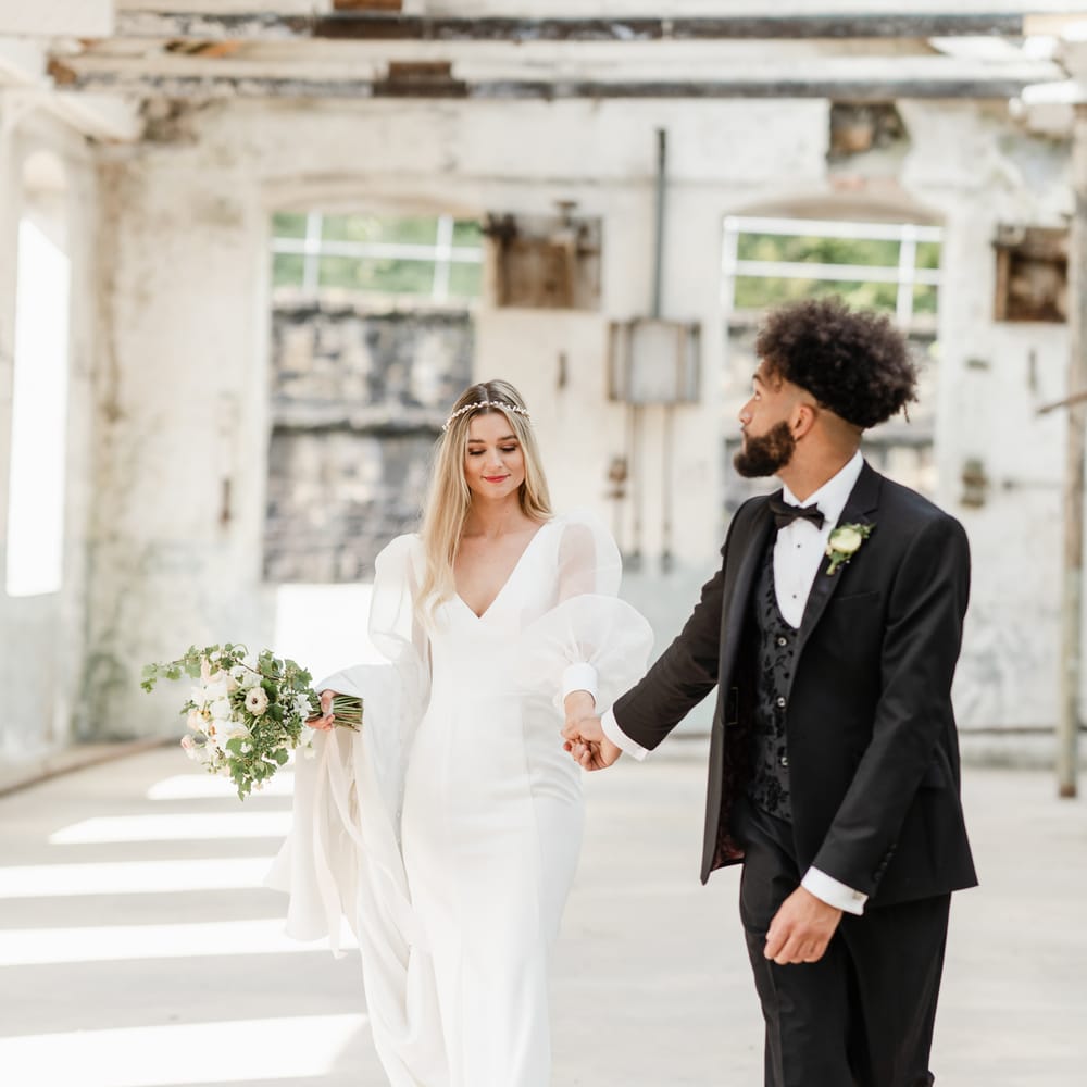 Groom in a black tuxedo and bow tie holding hands with his bride in a long sleeve wedding dress with halo headdress