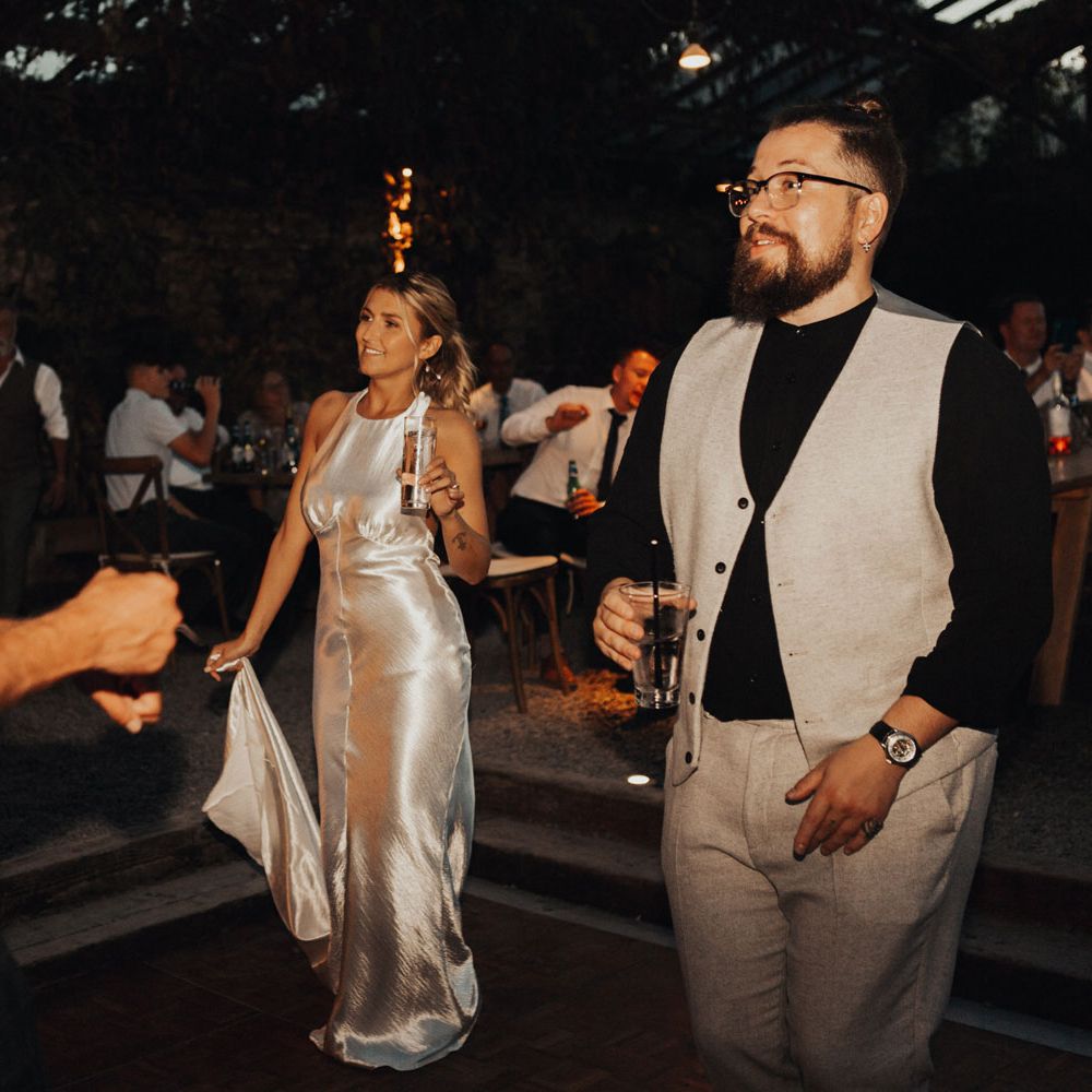 Bride with curled ponytail holds drink and train of her silver satin dress for wedding evening party at the glasshouse at Anran Devon