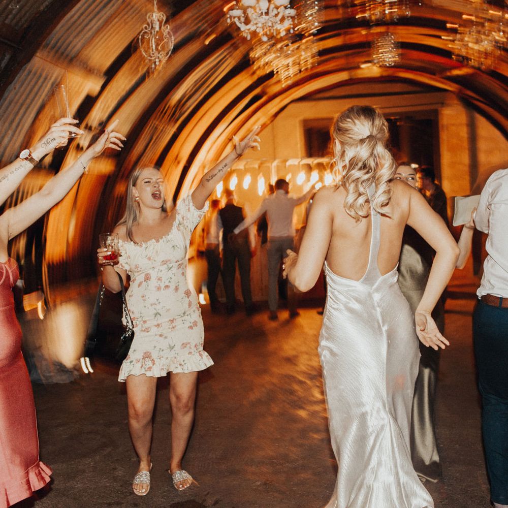Bride in racer back silver satin dress and curled blonde ponytail dances with wedding guests in the hut lit by chandeliers at Anran Devon