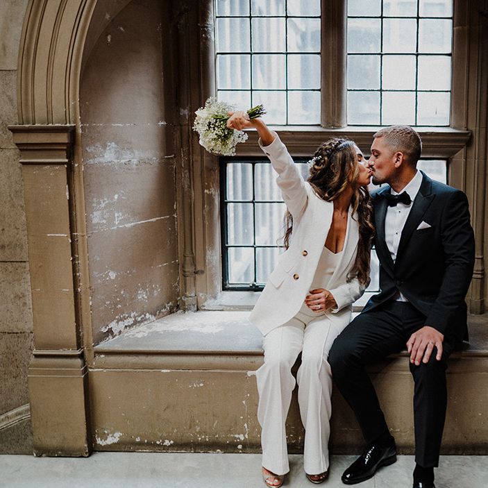 Groom in a black tuxedo kissing his bride in a white suit holding a white rose and gypsophila bouquet 
