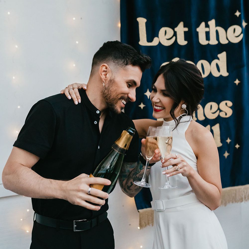 Bride in a halter neck wedding dress with belt detail and front split sipping champagne with her groom in a black shirt 