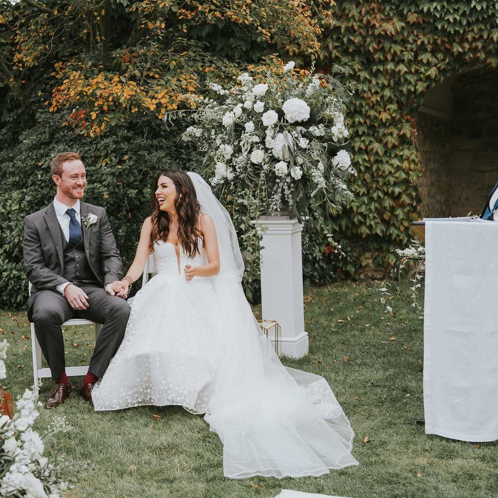 Bride in Pronovias wedding dress, Jimmy Choo wedding shoes and veil sits on white wooden chair during outdoor wedding ceremony with groom in grey three piece wedding suit, blue tie and white rose buttonhole