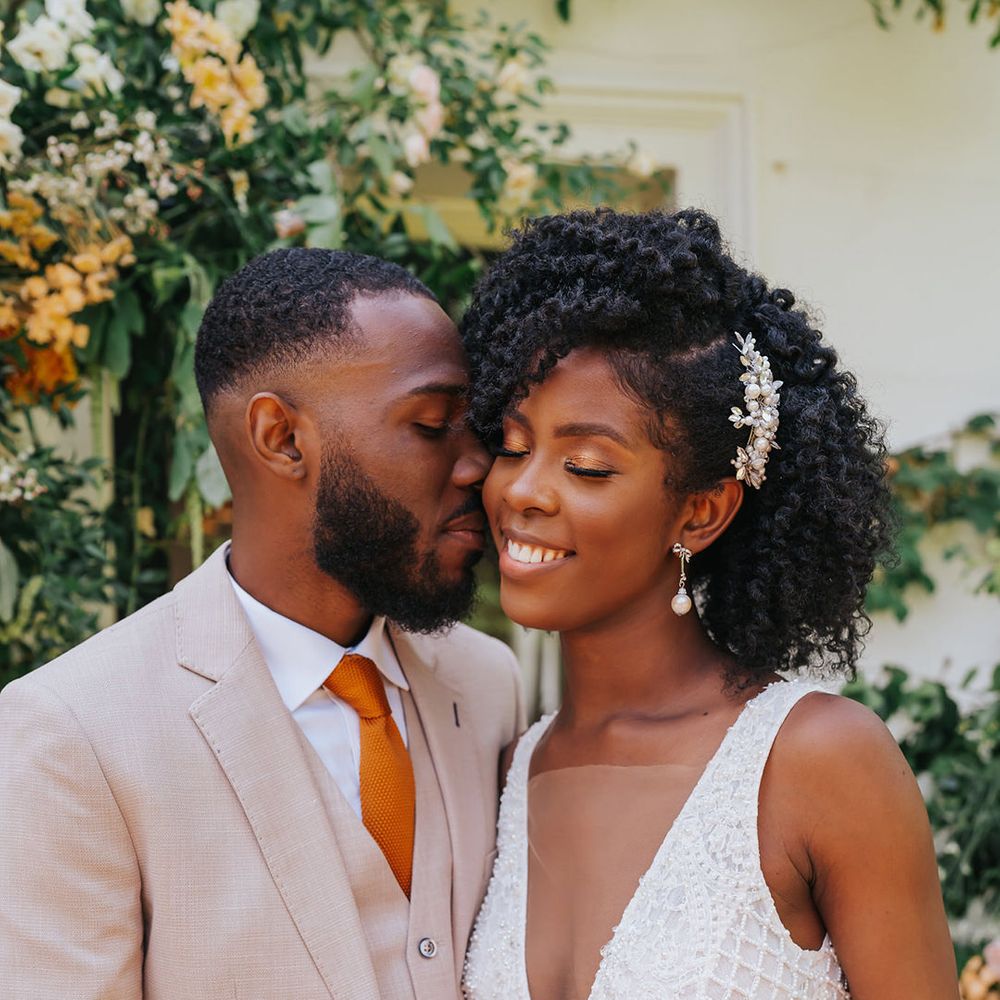 Black groom in a beige three-piece suit and orange tie kissing his bride in a plunging neckline wedding dress with naturally curly hair swept to one side and pinned with a jewel hair accessory 